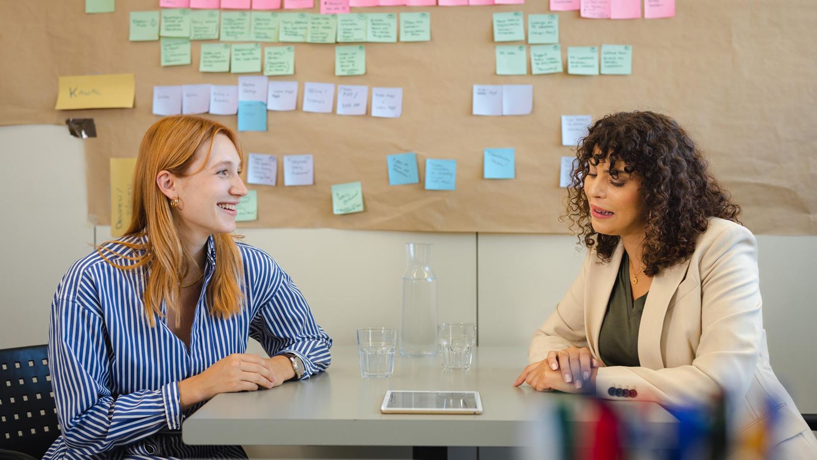 Two women sitting at a table having a conversation; a pinboard full of colourful notes hangs in the background.