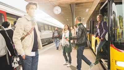 Pendlerinnen- und Pendler steigen an einem Bahnhof von einem Postauto auf ein Bahngleis um. Auf dem Bahngleis ist ein Zug der SBB zu sehen.   