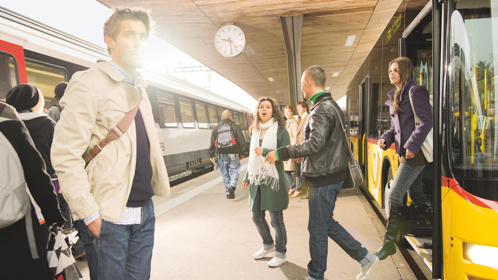 Pendlerinnen- und Pendler steigen an einem Bahnhof von einem Postauto auf ein Bahngleis um. Auf dem Bahngleis ist ein Zug der SBB zu sehen.