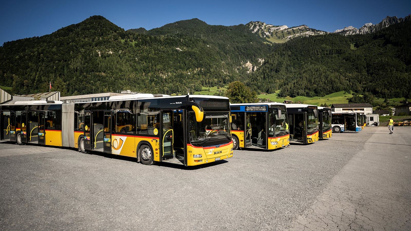 Fünf gelbe Postautos mit schwarzem Dach stehen nebeneinander auf einem Parkplatz inmitten einer alpinen Landschaft mit grünen Hügeln und felsigen Bergen unter blauem Himmel.