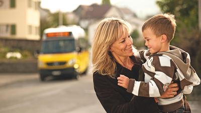 A woman has her son in her arms. A Postbus can be seen in the background.