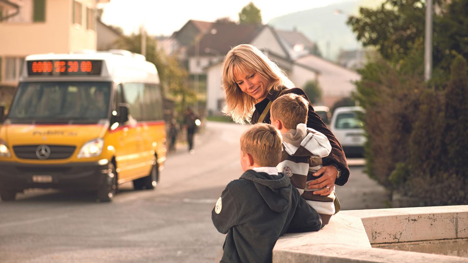 Una mamma aspetta con due bambini alla fermata di AutoPostale. Sullo sfondo è raffigurato un autopostale.