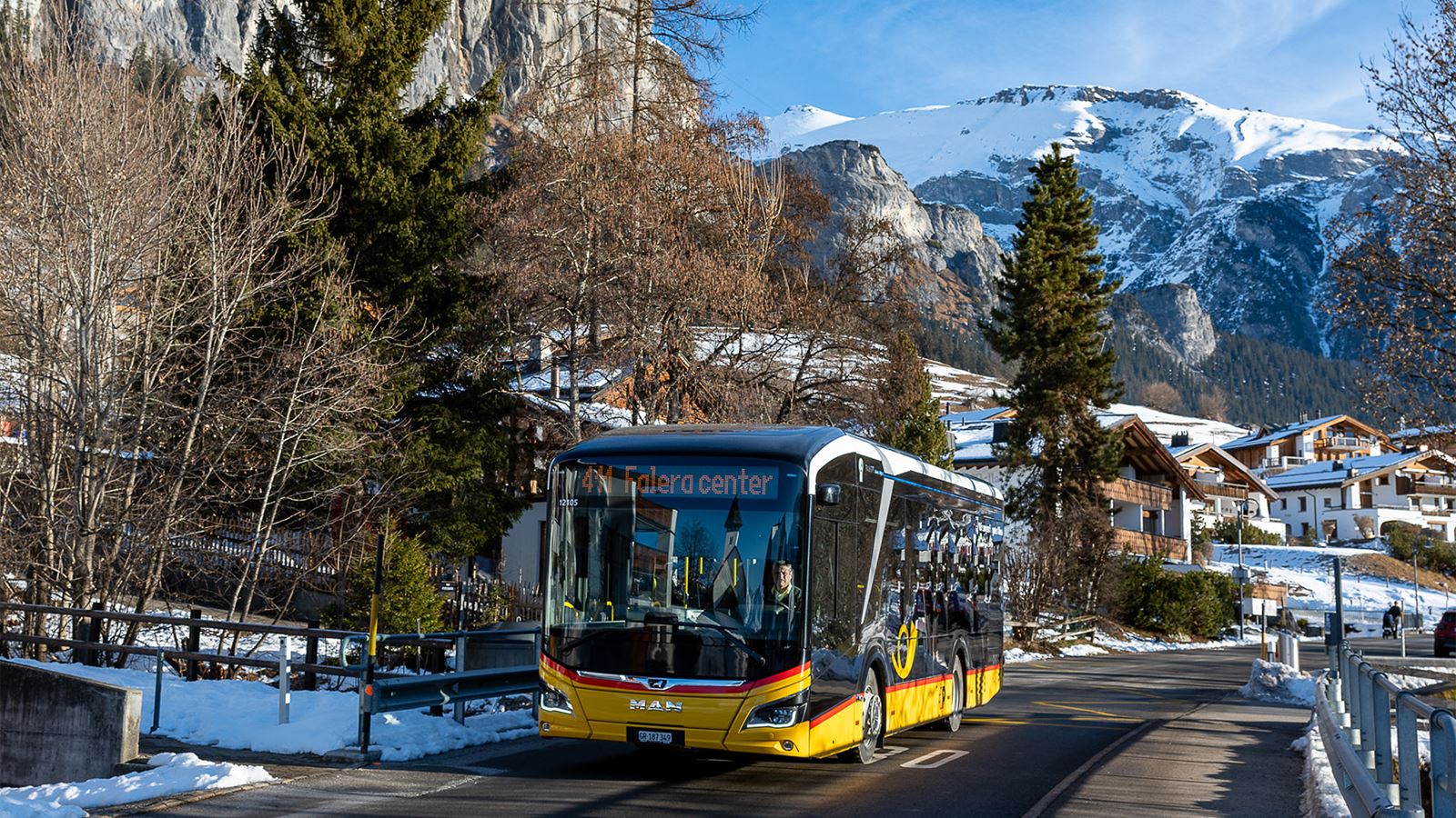 Das Postauto in einer verschneiten Schweizer Alpenlandschaft fährt durch ein Bergdorf mit traditionellen Holzhäusern und Blick auf hohe schneebedeckte Berge im Winter.