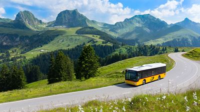 Postauto fährt auf Strasse durch Hügellandschaft. Im Hintergrund die Gantrisch-Bergkette. 