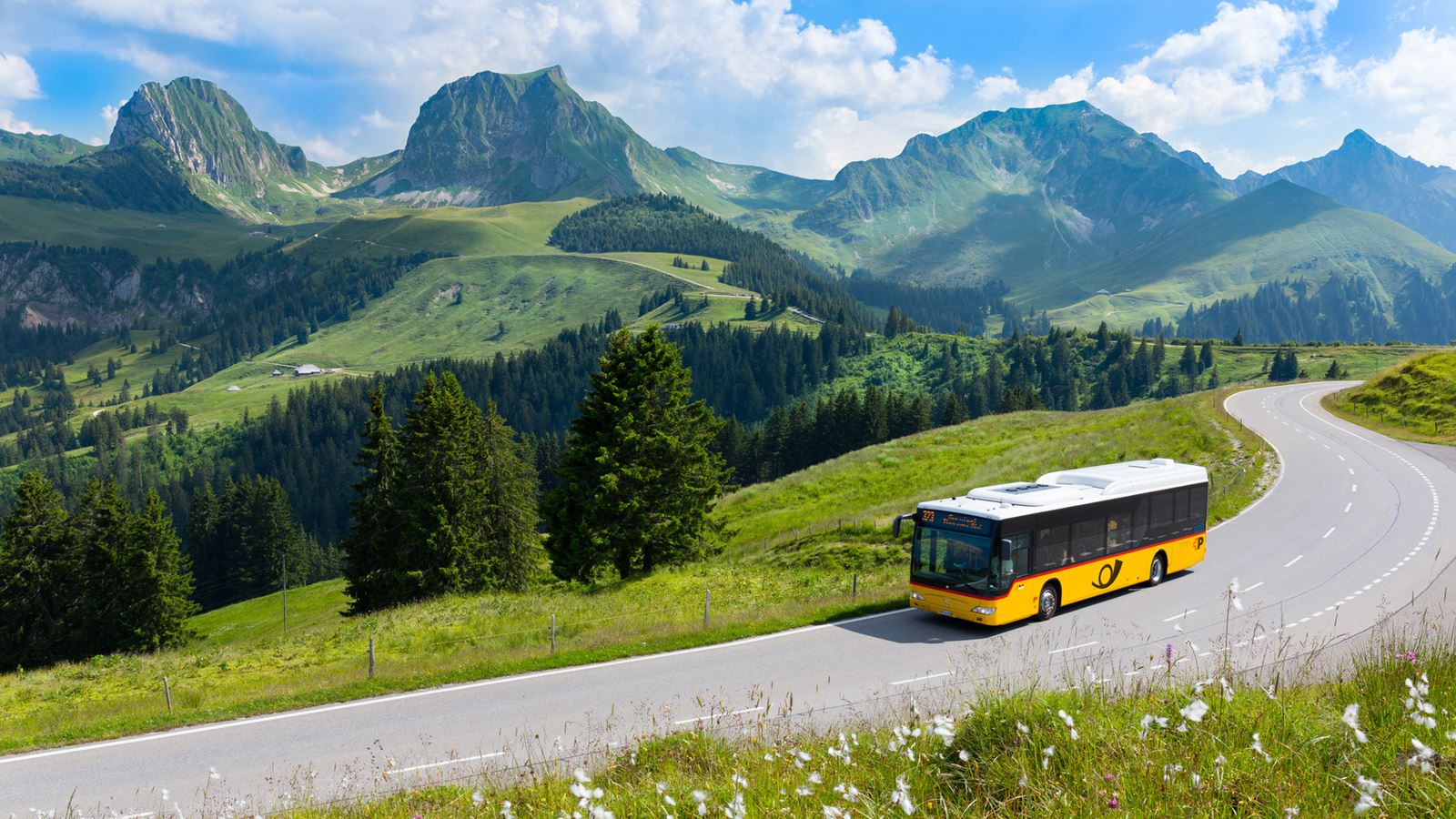 Postauto fährt auf Strasse durch Hügellandschaft. Im Hintergrund die Gantrisch-Bergkette.