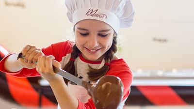 A girl in a Maestrani chef’s hat pours liquid chocolate into a pot with a ladle. © Maestrani Swiss Chocolates Ltd