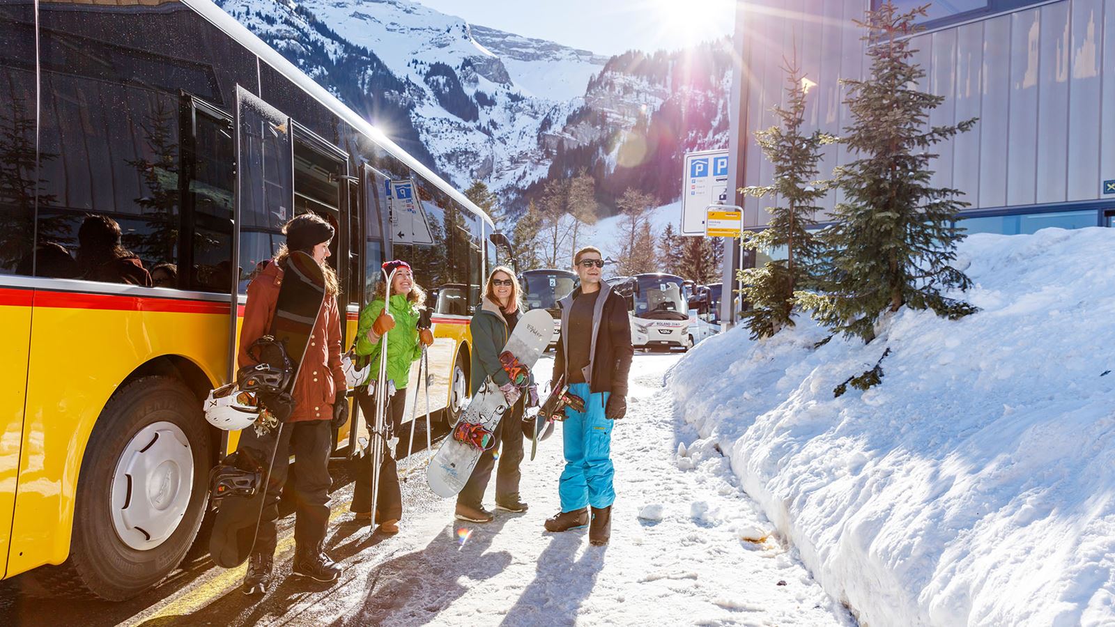 Four people get out of a Postbus in winter sports clothing with skis and snowboards.