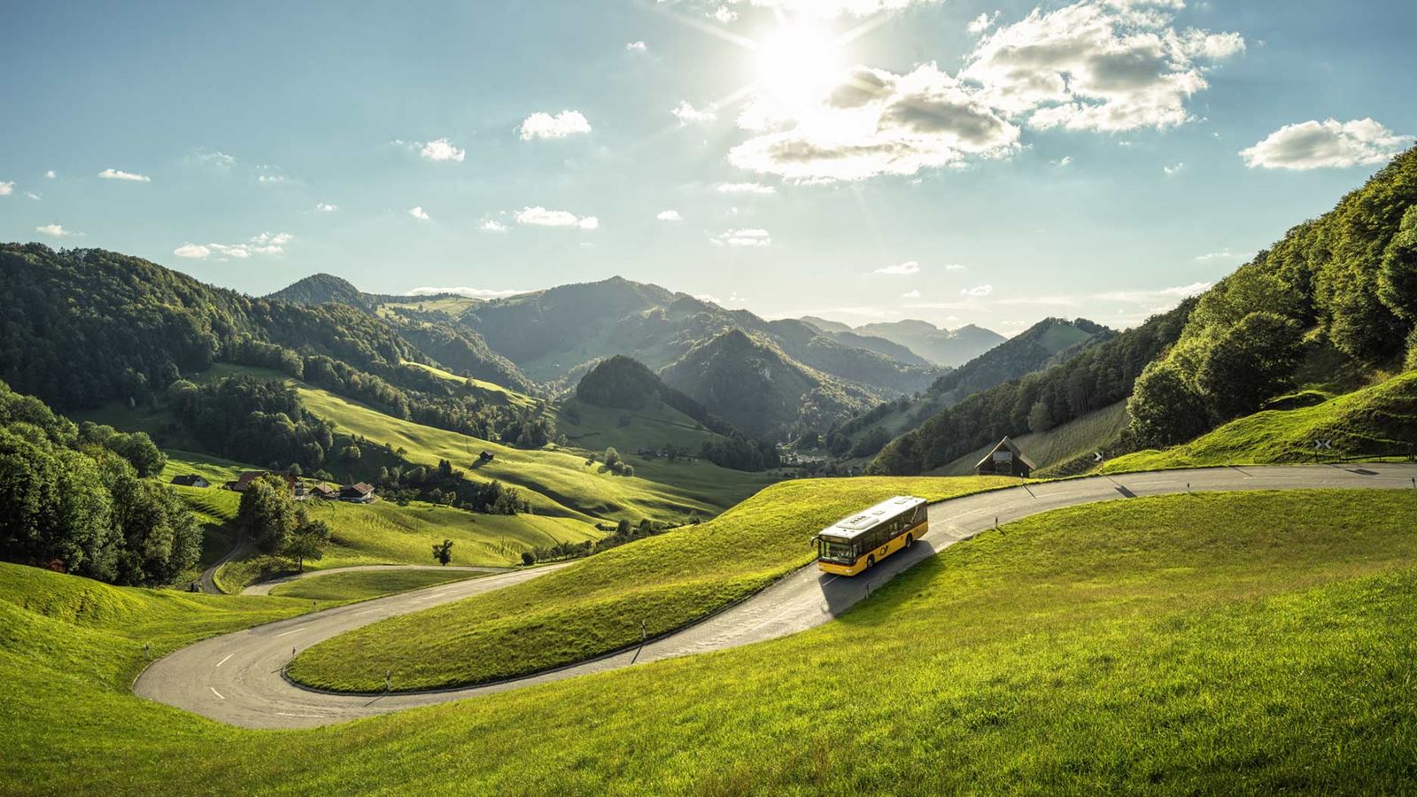 The Postbus drives down the passwang. The green, hilly landscape can be seen in the background.