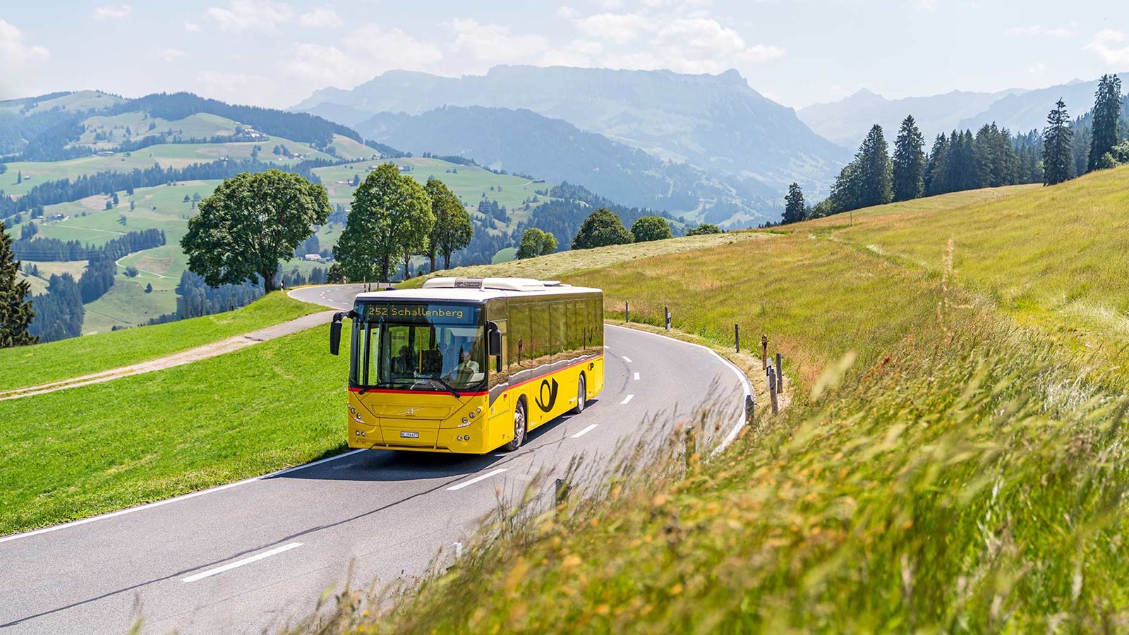 PostBus drives on roads in a rural area. In the background, the hilly landscape of Entlebuch with a view of the Schrattenfluh.