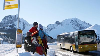 Fünfköpfige Familie mit Schneeschuhen wartet an der PostAuto-Station Wasserscheide auf das heranfahrende Postauto der Linie 323. Im Hintergrund Teil der verschneiten Gantrischkette.  