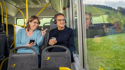 Eine Frau und ein Mann hören im Postauto das Hörerlebnis Umweg am Albis. Sie tragen Kopfhörer und schauen aus dem Fenster.