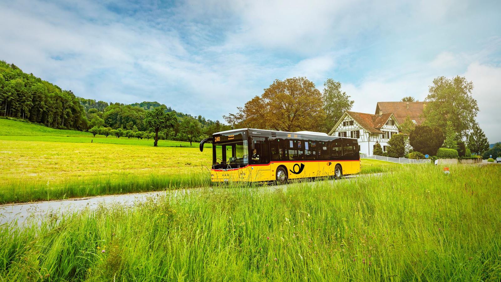 Ein gelbes Postauto fährt im Sommer durch ländliches Gebiet. Im Hintergrund ist ein traditionelles Riegelhaus zu sehen.