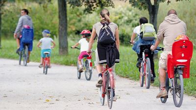 Erwachsene Personen und Kinder fahren mit dem Velo entlang grüner Bäume um den Flughafen Zürich.