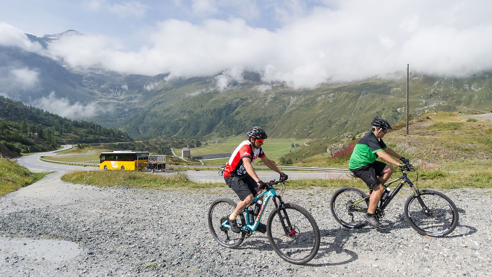 Zwei Mountainbiker auf dem Simplonpass. Im Hintergrund ist ein Postauto