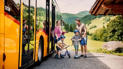 Zwei Erwachsene und zwei Kinder mit Helmen steigen neben dem Postauto auf einer Landstrasse in einer grünen Hügellandschaft aus.