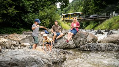Eine Familie rastet am Bach zwischen grossen Felsblöcken: Zwei Jungen spielen am Ufer, der Vater hockt und lässt Steine übers Wasser springen, die Mutter sitzt lächelnd auf einem Felsen. Im Hintergrund fährt ein Postauto über eine Brücke; ringsum dichter Wald und Hügellandschaft, sommerliches Licht.
