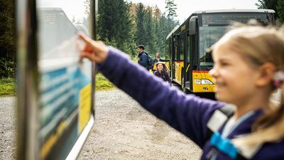Ein Kind schaut an einer Postauto Haltestelle den Fahrplan an, im Hintergrund steht ein Postauto.