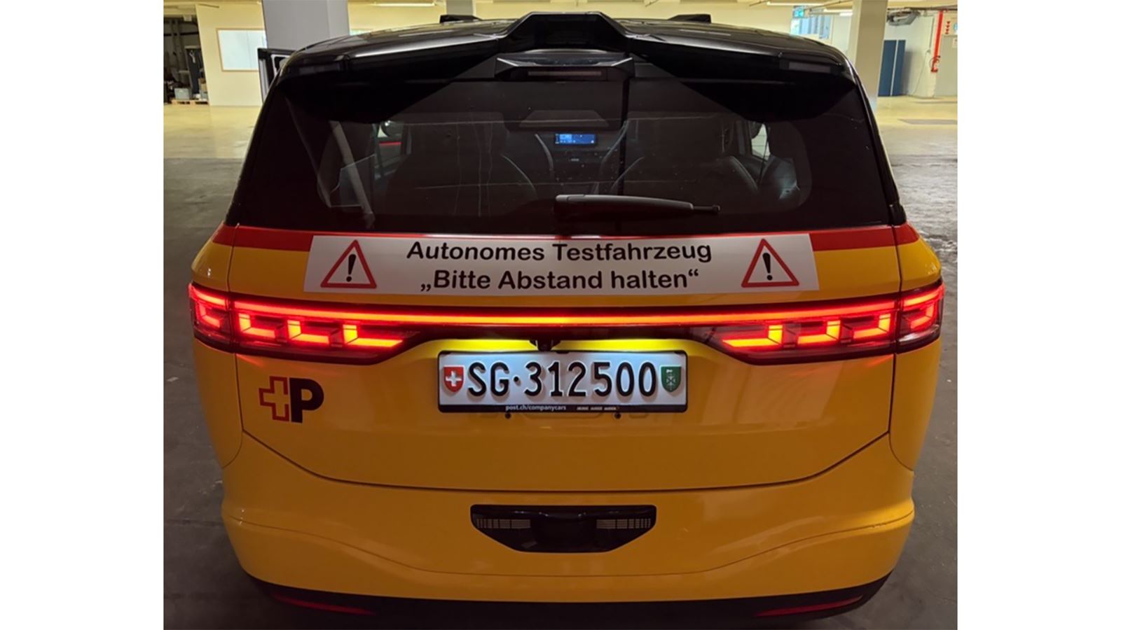 Un véhicule AmiGo jaune vu de derrière. Au-dessus de la plaque d’immatriculation se trouve une affiche comportant l’inscription «Véhicule d’essai autonome, veuillez garder vos distances!». 