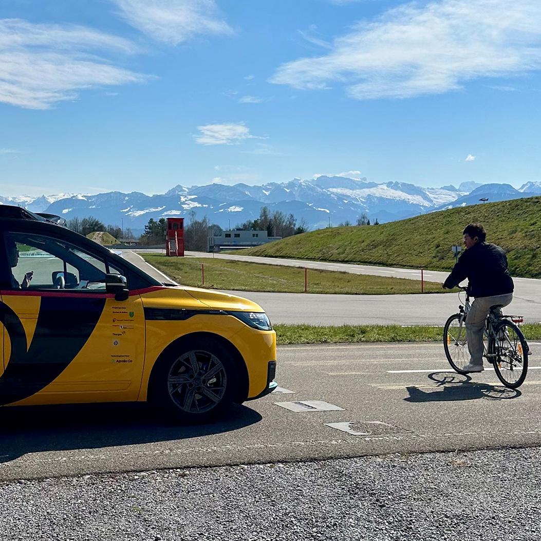 Gelbes AmiGo Fahrzeug hält auf Teststrecke an, während eine Person mit Velo die Strasse überquert; im Hintergrund sind Berge zu sehen.