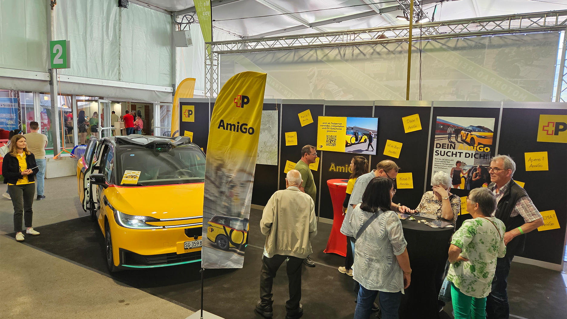 Gelbes AmiGo-Fahrzeug am Messestand steht mit geöffneten Türen in einer Halle; im Hintergrund Informationswände und Besuchende.