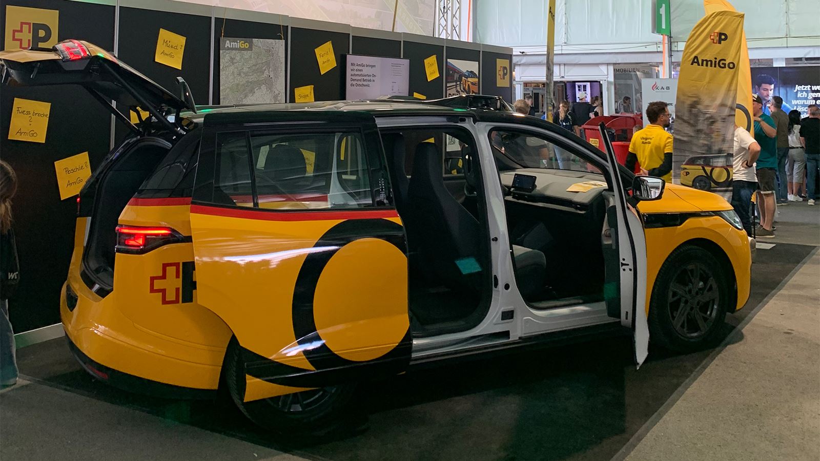 Un veicolo AmiGo giallo allo stand fieristico si trova in un padiglione con le portiere aperte; sullo sfondo bacheche informative e visitatori.