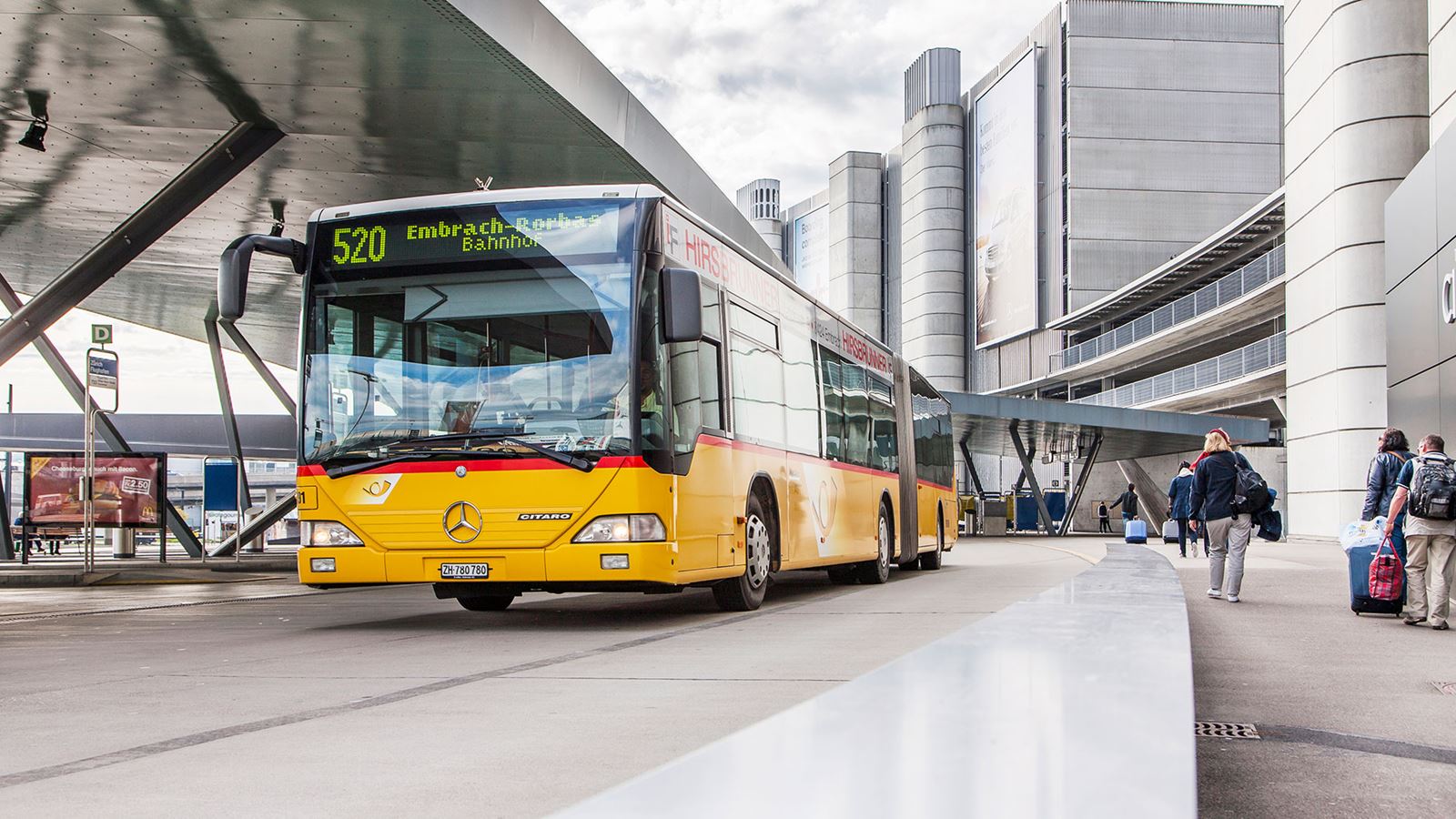 Ein Postauto hält an einem Bahnhof in einem städtischen Umfeld.