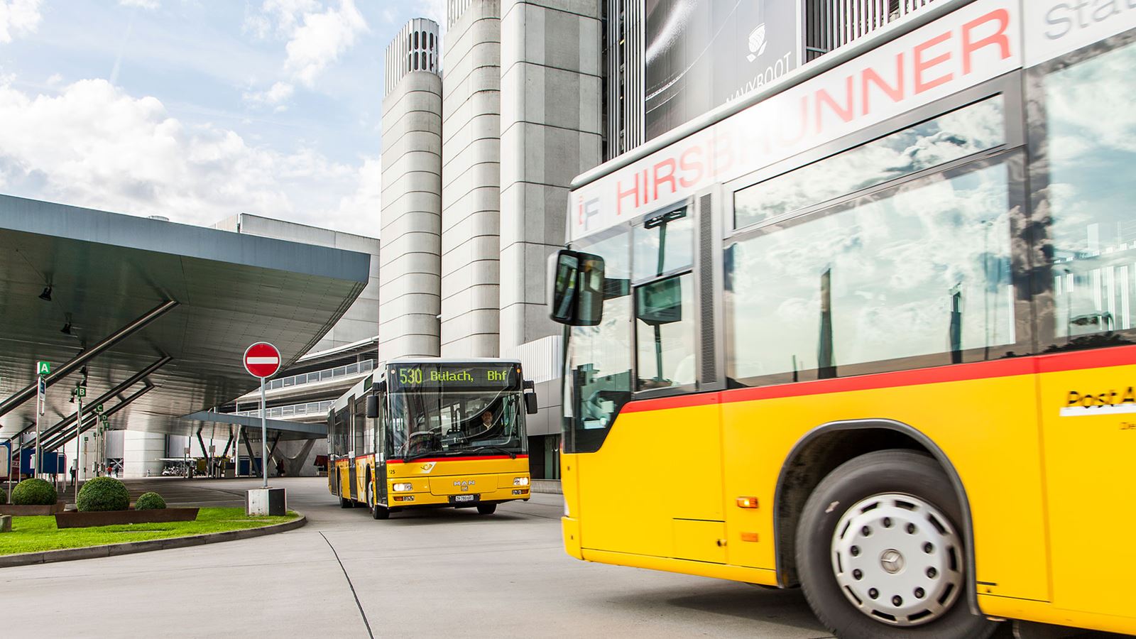 Zwei Postautos kreuzen sich an einer Haltestelle vor einem Hochhaus.