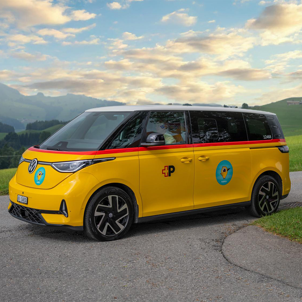Electric VW bus in the yellow PostBus design with red stripe and post horn logo, used for the Publicar on-demand service in Appenzell. The vehicle is standing on a country road in picturesque Swiss hilly landscape, next to a sign for hiking and cycling trails