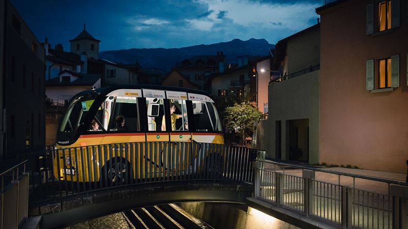 The PostBus SmartShuttle travelling across a small bridge in Sion at night. 