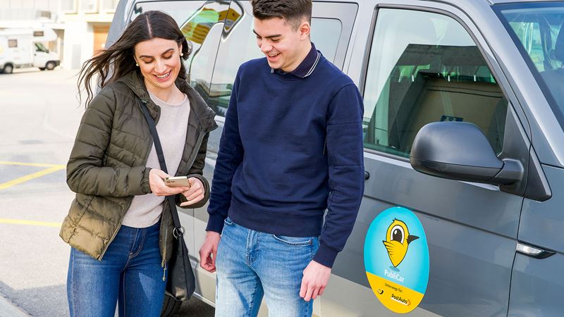 Photo of two people standing in front of a grey Kollibri taxi and looking at a smartphone. 