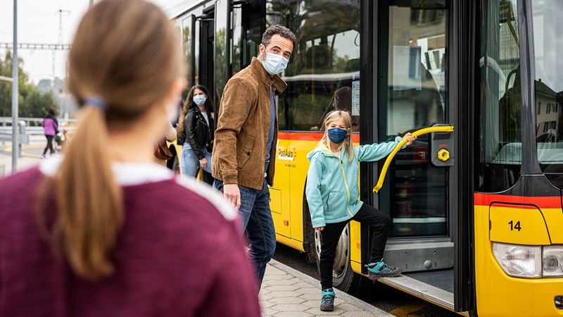 Ein erwachsener Mann und ein Kind welche ins Postauto einsteigen. Beide tragen Masken über ihren Gesichtern. 