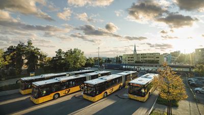Mehrere Postautos stehen auf einem Parkplatz, im Hintergrund sind Bäume, Gebäude und ein Zug zu sehen. 