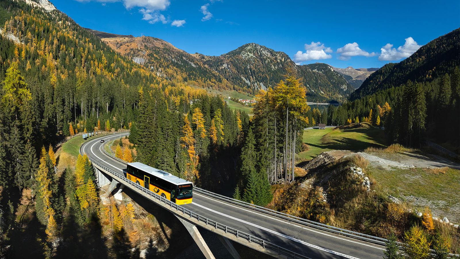Gelber Postauto-Bus fährt über eine Brücke durch eine herbstliche Berglandschaft mit dichten Wäldern, kurviger Straße und bewaldeten Gipfeln unter blauem Himmel.